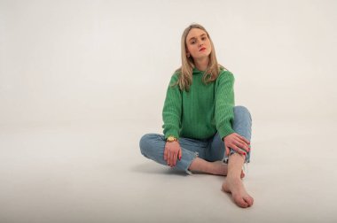 Young, cute, emotional, chic girl sitting on the floor in a flirtatious mood, hand gestures, empty space, wearing jeans and a green sweater on an isolated white background, place for text