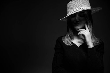 Black and white photo, portrait of a young, beautiful girl in a black dress with a straw hat on a gray background. Emotions and modesty looking down. Low tone