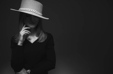 Black and white photo, portrait of a young, beautiful girl in a black dress with a straw hat on a gray background. Emotions and modesty looking down. Low tone