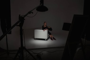 Sahne arkasında fotoğraf çekimi ayarlanıyor: kız fotoğraf çekimi için hazırlanıyor, kanepede oturuyor, kamera ekipmanlarının etrafında. Moda dergisi fotoğraf stüdyosu, gösterişsiz, gösterişsiz.