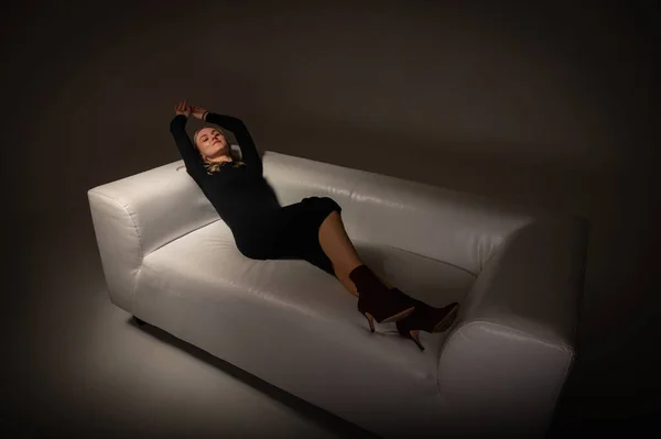 黒のドレスを着た若いかなり感情的な女の子がソファの上に横になって疲れ目を上げてリラックス光の背景に光の線 — ストック写真