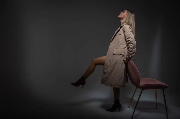 穿着灰色雨衣的年轻 情绪化的女孩站在椅子旁边 抬起一条腿 靠着白色的背景 闪烁着光芒 — 图库照片