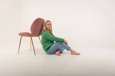 portrait of a young emotional girl sitting on the floor near a chair in a flirtatious mood, gesturing with her hands, empty space, wearing jeans and a green sweater n, space for text