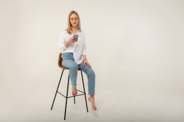Kot pantolonlu, beyaz gömlekli, cep telefonlu, beyaz arka planda izole edilmiş bir taburede oturan çekici bir kızın tam boy yan profil fotoğrafı.