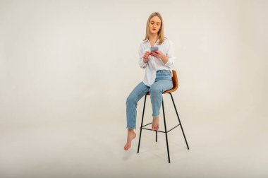 Kot pantolonlu, beyaz gömlekli, cep telefonlu, beyaz arka planda izole edilmiş bir taburede oturan çekici bir kızın tam boy yan profil fotoğrafı.