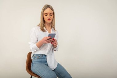 Full length side profile photo of young attractive girl in jeans and white shirt looking, browsing mobile phone while sitting on stool isolated on white background