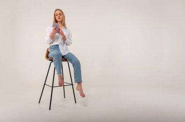 Full length side profile photo of young attractive girl in jeans and white shirt looking, browsing mobile phone while sitting on stool isolated on white background