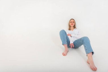 Portrait of a smiling young pretty European woman relaxing while sitting on the floor in jeans and a white shirt, isolated on white background. Place for writing