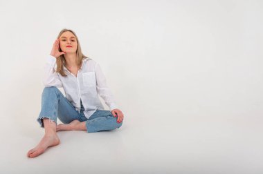 Portrait of a smiling young pretty European woman relaxing while sitting on the floor in jeans and a white shirt, isolated on white background. Place for writing