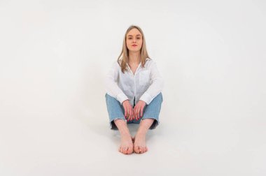 Portrait of a smiling young pretty European woman relaxing while sitting on the floor in jeans and a white shirt, isolated on white background. Place for writing