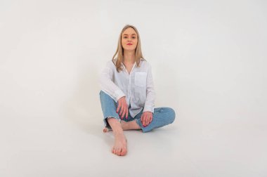 Portrait of a smiling young pretty European woman relaxing while sitting on the floor in jeans and a white shirt, isolated on white background. Place for writing
