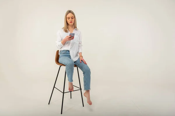 全长侧写照片 年轻迷人的女孩穿着牛仔裤和白衬衫 坐在白色背景的凳子上 看起来像在浏览手机 — 图库照片