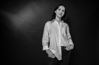 Siyah beyaz iş portresi, yakın plan güzel bir kız, duygusal dalgın bir gülümseme, koyu arka plan, beyaz gömlek ve kot pantolon, kollar çapraz, fotokopi alanı ile çekilen stüdyo fotoğrafı.