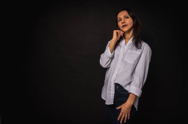 Genç bir kızın portresi, tatlı, duygusal dalgın, koyu arkaplanda duran, beyaz gömlek ve kot pantolon, el ele, fotokopi ile çekilen stüdyo fotoğrafı.