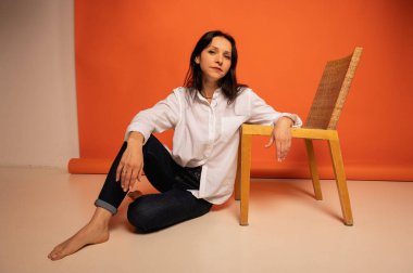 Çekici, şık bir iş kadınının sandalyede otururken çekilmiş uzun bir fotoğrafı. Flört havası, kot pantolonlu boş bir yer ve turuncu arka planda beyaz gömlek. Mesaj için yer.