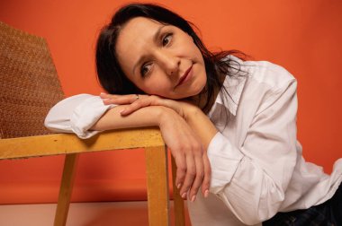 Çekici, şık bir iş kadınının sandalyede otururken çekilmiş uzun bir fotoğrafı. Flört havası, kot pantolonlu boş bir yer ve turuncu arka planda beyaz gömlek. Mesaj için yer.