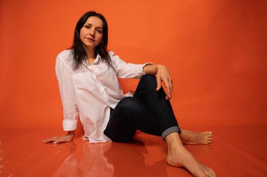 40 yaşlarında, tatlı, duygusal olarak düşünceli, turuncu bir arka planda yerde oturan, elleri kapalı, kapalı, fotokopi makinesiyle çekilen genç bir kızın iş portresi.