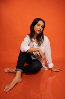 40 yaşlarında, tatlı, duygusal olarak düşünceli, turuncu bir arka planda yerde oturan, elleri kapalı, kapalı, fotokopi makinesiyle çekilen genç bir kızın iş portresi.