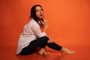 40 yaşlarında, tatlı, duygusal olarak düşünceli, turuncu bir arka planda yerde oturan, elleri kapalı, kapalı, fotokopi makinesiyle çekilen genç bir kızın iş portresi.