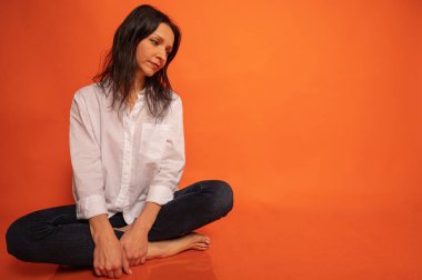 40 yaşlarında, tatlı, duygusal olarak düşünceli, turuncu bir arka planda yerde oturan, elleri kapalı, kapalı, fotokopi makinesiyle çekilen genç bir kızın iş portresi.