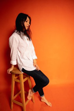 40 yaşlarında genç ve güzel bir kızın iş portresi. Tahta bir sandalyede, parlak turuncu bir arka planda oturuyor. Elleriyle işaret ediyor.