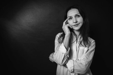 Siyah beyaz iş portresi, yakın plan güzel bir kız, duygusal dalgın bir gülümseme, koyu arka plan, beyaz gömlek ve kot pantolon, kollar çapraz, fotokopi alanı ile çekilen stüdyo fotoğrafı.