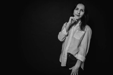 Siyah beyaz iş portresi, yakın plan güzel bir kız, duygusal dalgın bir gülümseme, koyu arka plan, beyaz gömlek ve kot pantolon, kollar çapraz, fotokopi alanı ile çekilen stüdyo fotoğrafı.