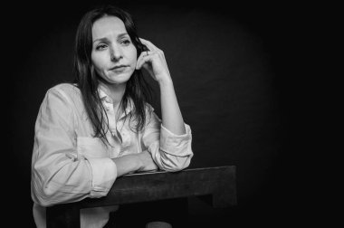 Siyah-beyaz, genç güzel bir kızın iş portresi, duygusal düşünceli gülümseme, koyu arka planda durmak, beyaz gömlek ve kot pantolon, kollar çapraz, fotokopi ile çekilen stüdyo fotoğrafı.