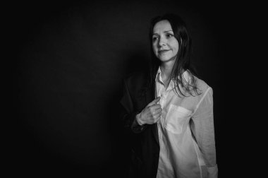 Siyah-beyaz, genç güzel bir kızın iş portresi, duygusal düşünceli gülümseme, koyu arka planda durmak, beyaz gömlek ve kot pantolon, kollar çapraz, fotokopi ile çekilen stüdyo fotoğrafı.