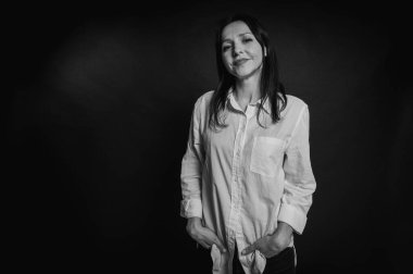 Genç ve güzel bir kızın iş portresi, duygusal düşünceli gülümseme, koyu arka planda durmak, beyaz gömlek ve kot pantolon, kollar çapraz, fotokopi ile çekilen stüdyo fotoğrafı.