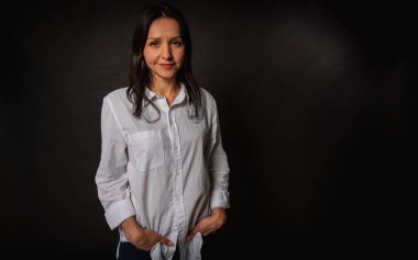 Genç ve güzel bir kızın iş portresi, duygusal düşünceli gülümseme, koyu arka planda durmak, beyaz gömlek ve kot pantolon, kollar çapraz, fotokopi ile çekilen stüdyo fotoğrafı.
