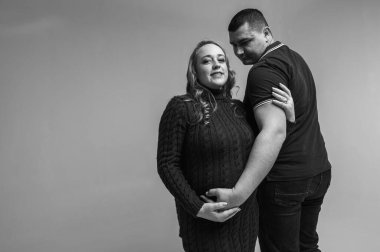 Beyaz bir duvarda izole edilmiş hamile karısıyla ayakta duran mutlu genç bir adamın siyah beyaz görüntüsü. Aile büyümesi, çocuk beklemesi, genç aile, duygular.