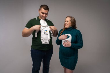 Genç, neşeli, gülümseyen bir baba ve ellerinde bebek tulumlarıyla hamile bir anne. Bir çocuk bekliyorum.