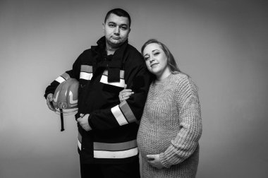 İtfaiyeci kocasıyla birlikte siyah-beyaz, hamile bir kız arka planda izole edilmiş itfaiyeci üniformasıyla karnını elleriyle destekleyen bir çocuk bekliyor. Bir çocuk bekliyorum.