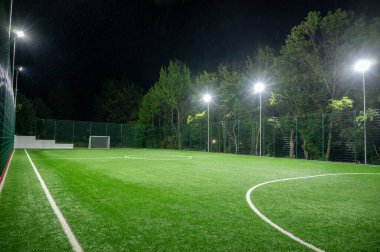 Parktaki spor sahasında yeşil ağaçların arkasında yapay çimen stadyumu var. Güçlü fenerlerle gece aydınlatması, aşağıdan manzara