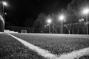 Siyah beyaz fotoğraf, yeşil ağaçların arka planında stadyumun yapay çimleriyle parkta akşam sporu sahası. Güçlü fenerlerle gece aydınlatması, aşağıdan manzara