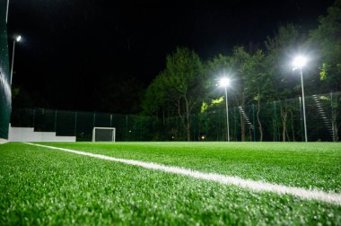 Parktaki spor sahasında yeşil ağaçların arkasında yapay çimen stadyumu var. Güçlü fenerlerle gece aydınlatması, aşağıdan manzara
