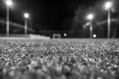 Siyah beyaz fotoğraf, yeşil ağaçların arka planında stadyumun yapay çimleriyle parkta akşam sporu sahası. Güçlü fenerlerle gece aydınlatması, aşağıdan manzara