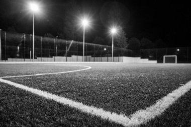 Siyah beyaz fotoğraf, yeşil ağaçların arka planında stadyumun yapay çimleriyle parkta akşam sporu sahası. Güçlü fenerlerle gece aydınlatması, aşağıdan manzara