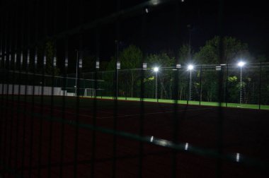 Parktaki spor sahasında yeşil ağaçların arkasında yapay çimen stadyumu var. Güçlü fenerlerle gece aydınlatması, aşağıdan manzara, sade anahtar.