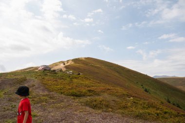 Gökyüzü Ukrayna dağlarının üzerinde mavi, kırmızı tişörtlü ve siyah şapkalı küçük bir çocuk dağlara tırmanıyor. Dağlarda dinlen.