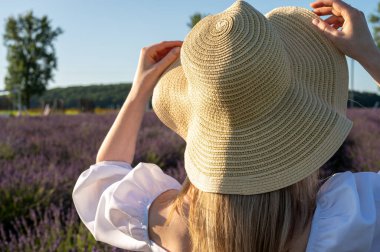 Lavanta tarlasında gün batımında hasır şapkalı sarışın bir kız elini ve beyaz elbisesini düzeltir. Arka görünüm