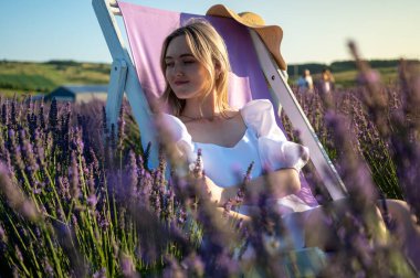 Beyaz elbiseli rahat genç bir kız, bir sandalyede oturuyor, temiz hava soluyor, güneşli bir günde lavanta tarlasında oturuyor, yan tarafa bakıyor, düşünüyor