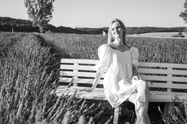 Siyah ve beyaz fotoğraf, beyaz elbiseli rahat bir genç kızın portresi, bir sandalyede oturuyor, temiz hava soluyor, güneşli bir günde lavanta tarlasında oturuyor, başka yere bakıyor,