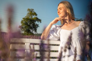 Beyaz elbiseli rahat bir genç kızın portresi, beyaz bir bankta oturuyor, temiz hava soluyor, güneşli bir günde lavanta tarlasında oturuyor, başka yere bakıyor, düşünüyor.