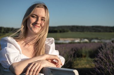 Beyaz elbiseli rahat bir genç kızın portresi, beyaz bir bankta oturuyor, temiz hava soluyor, güneşli bir günde lavanta tarlasında oturuyor, başka yere bakıyor, düşünüyor.