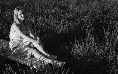 Siyah ve beyaz fotoğraf, beyaz bir bankta oturan, beyaz elbiseli rahat bir genç kızın portresi, temiz hava soluyor, güneşli bir günde lavanta tarlasında oturuyor, başka yere bakıyor, düşünüyor.