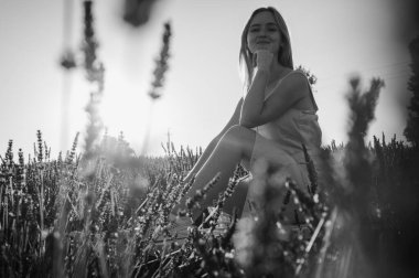 Günbatımının ışınlarında siyah beyaz bir portre, beyaz elbiseli rahat bir genç kızın yan portresi, beyaz bir bankta oturuyor, temiz hava soluyor, lavanta tarlasında oturuyor.