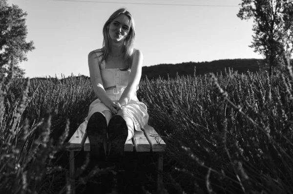 Siyah ve beyaz fotoğraf, beyaz bir bankta oturan, beyaz elbiseli rahat bir genç kızın portresi, temiz hava soluyor, güneşli bir günde lavanta tarlasında oturuyor, başka yere bakıyor, düşünüyor.