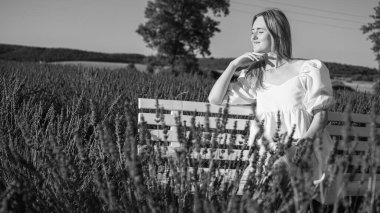 Siyah ve beyaz fotoğraf, beyaz elbiseli rahat bir genç kızın portresi, bir sandalyede oturuyor, temiz hava soluyor, güneşli bir günde lavanta tarlasında oturuyor, başka yere bakıyor,